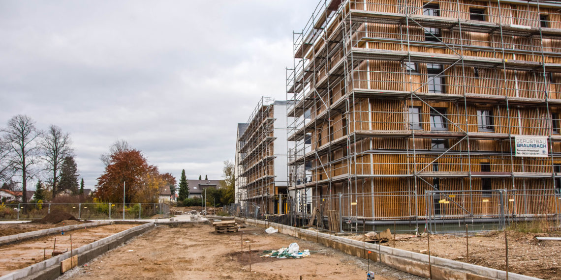 Das Bild zeigt eine Baustelle, auf der rechts ein Holzhaus mit Gerüst steht. Der Boden ist matschig, im Hintergrund steht ein Kran.