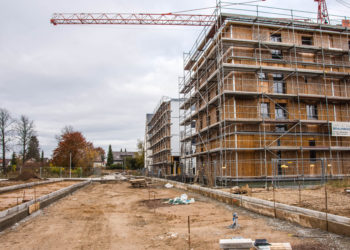 Das Bild zeigt eine Baustelle, auf der rechts ein Holzhaus mit Gerüst steht. Der Boden ist matschig, im Hintergrund steht ein Kran.