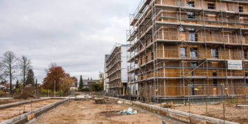 Das Bild zeigt eine Baustelle, auf der rechts ein Holzhaus mit Gerüst steht. Der Boden ist matschig, im Hintergrund steht ein Kran.
