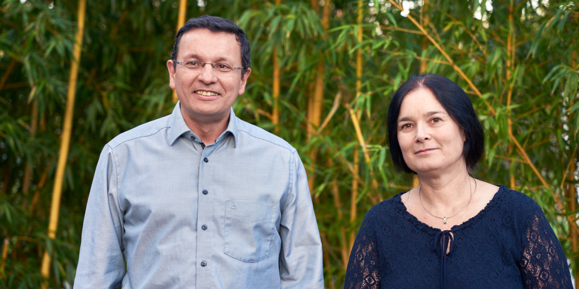 Eine Frau im blauen Oberteil und ein Mann im hellblauen Hemd lächeln in die Kamera. Sie stehen vor einem Bambus