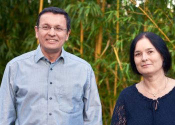 Eine Frau im blauen Oberteil und ein Mann im hellblauen Hemd lächeln in die Kamera. Sie stehen vor einem Bambus