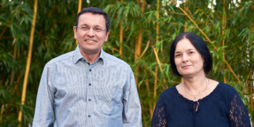 Eine Frau im blauen Oberteil und ein Mann im hellblauen Hemd lächeln in die Kamera. Sie stehen vor einem Bambus